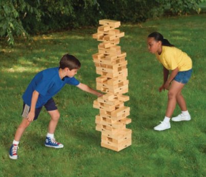 Two children joyfully playing with a giant wooden Jenga tower in a sunny yard.